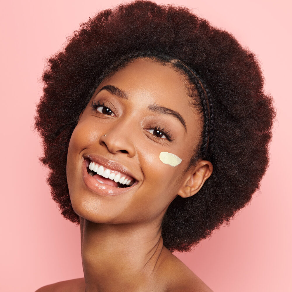 A black woman with a short afro smiles at the camera with a small amount of facial treatment applied to her left cheek.