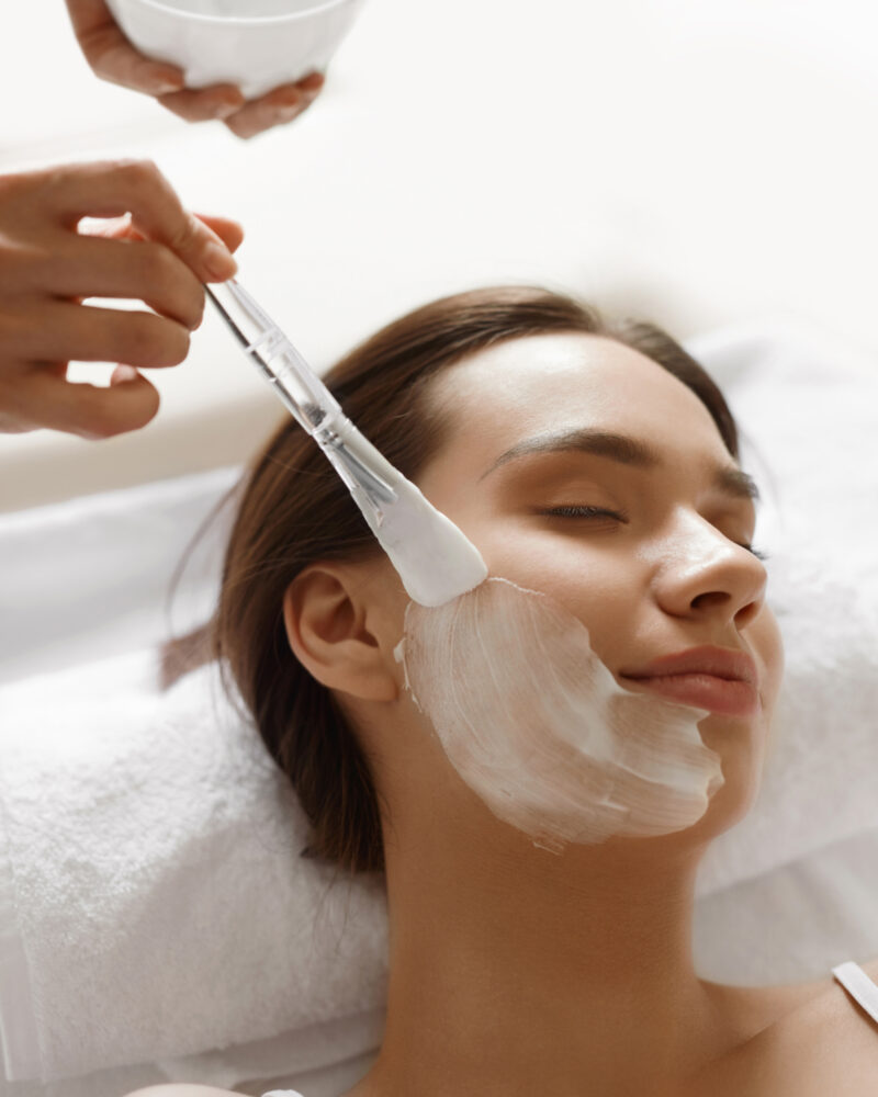 A technicians hands painting a facial treatment onto a white woman's face.