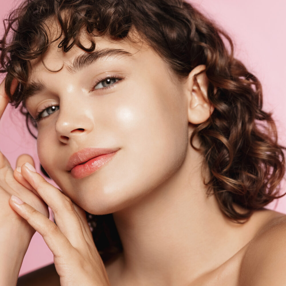 A white woman with curly brown hair and bangs rests her face on her hands while looking at the camera.