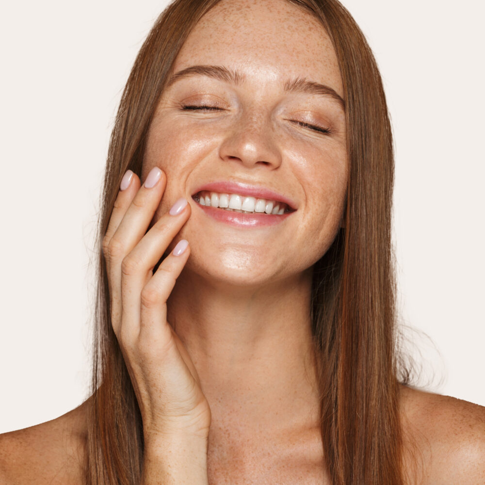 A white woman with long light brown hair smiles with her eyes closed and touches her face with her right hand.