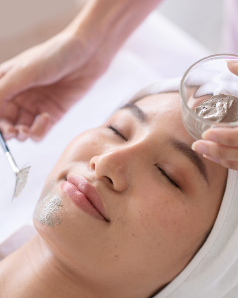 An Asian woman lies on a table with her hair covered in a towel and has a facial treatment applied with a brush.