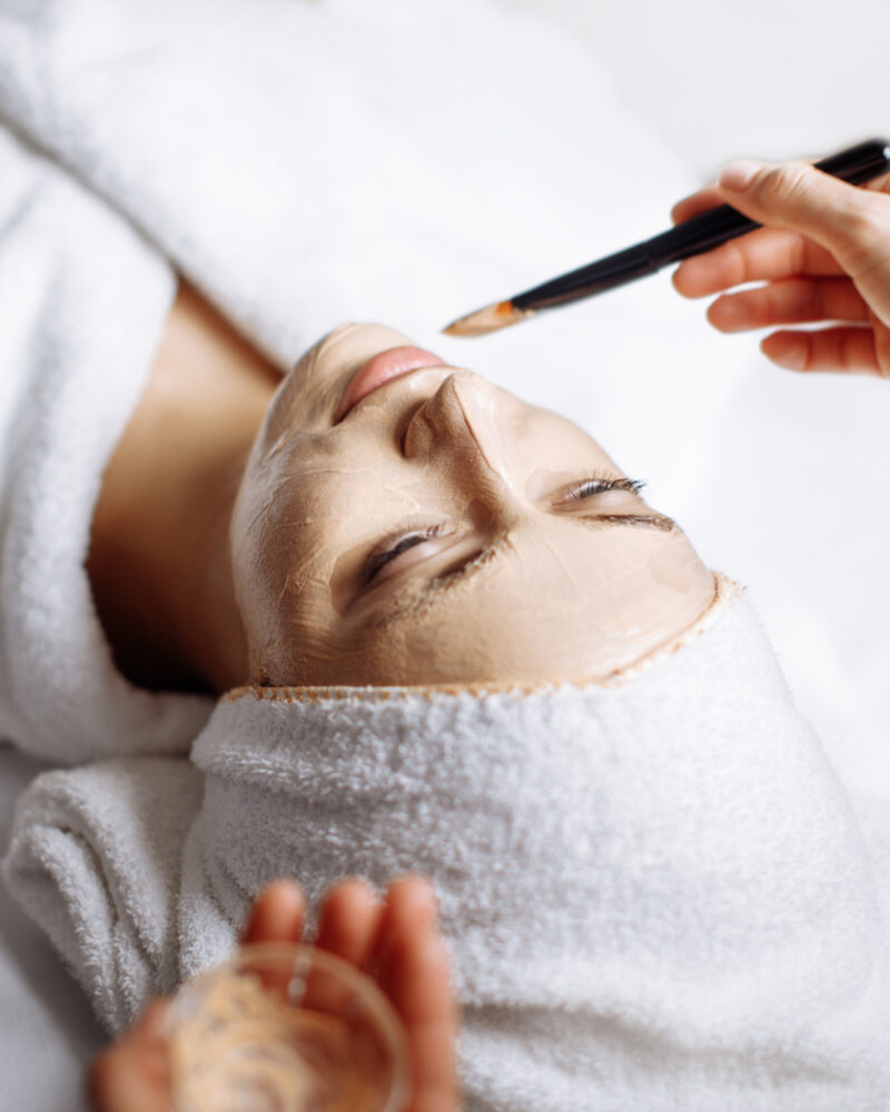 A technicians hand painting a facial treatment onto a white woman's face.